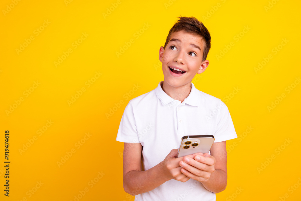 Photo of funky impressed shool boy dressed polo t-shirt working modern gadget looking emtpy space isolated yellow color background Photo of funky impressed shool boy dressed polo t-shirt working modern gadget looking emtpy space isolated yellow color background