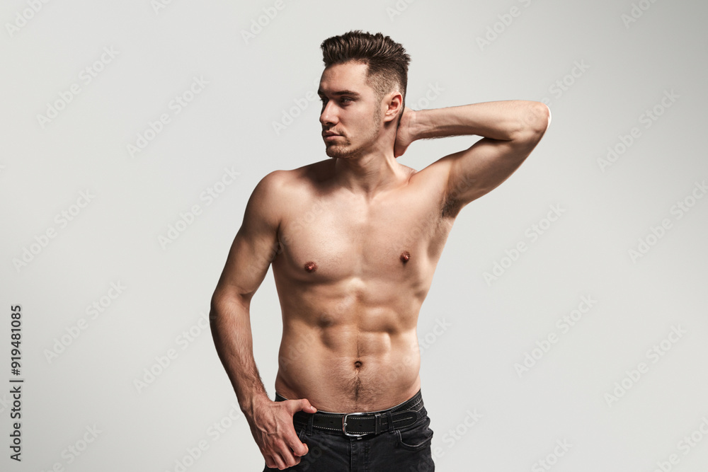 A muscular man posing shirtless in a studio with his hand on his neck, highlighting fitness and strength. A muscular man posing shirtless in a studio with his hand on his neck, highlighting fitness and strength.