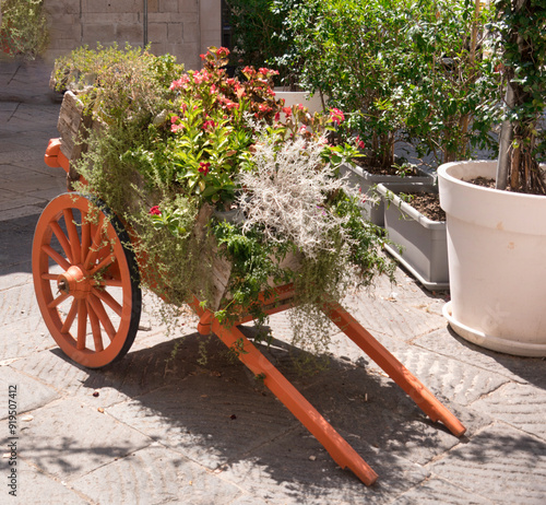 Rustic wheeled red cart in a stone square full bright colored flowers in Italy photo