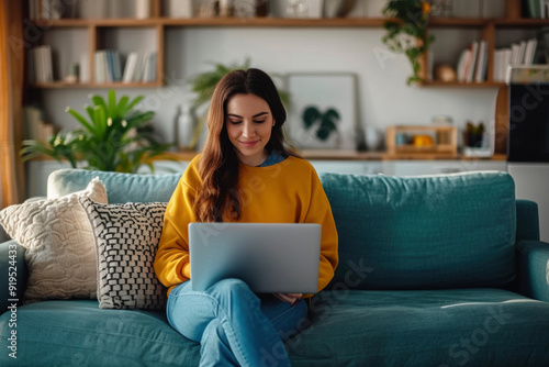 A young woman is sitting on a couch in her living room, working on her laptop. She is smiling and appears to be focused on her work.