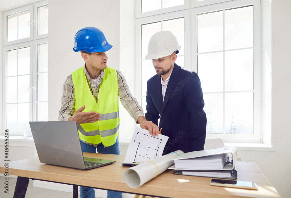 Two men in construction helmets discuss the master plan in a room, close-up. Concept design department. The inspector checks the construction plan, permitting documents, front view Two men in construction helmets discuss the master plan in a room, close-up. Concept design department. The inspector checks the construction plan, permitting documents, front view