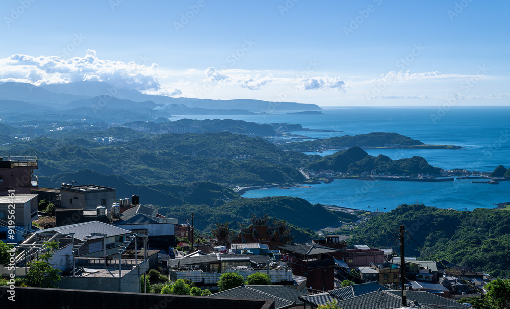 Fototapeta premium A scenic view of Jiufen Village with green mountains stretching to the blue sea, under a blue sky with white clouds. The picturesque afternoon landscape. Jiufen, Taiwan.
