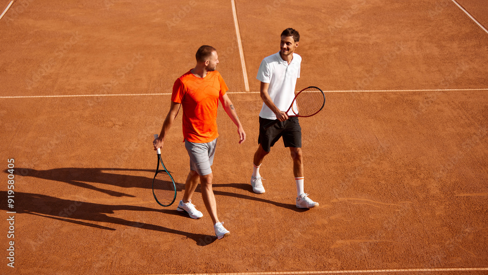 Fototapeta premium Two men, tennis players walking on outdoor clay court with racket, discussing strategy of the game. Team training and competing. Concept of sport, competition, active and healthy lifestyle