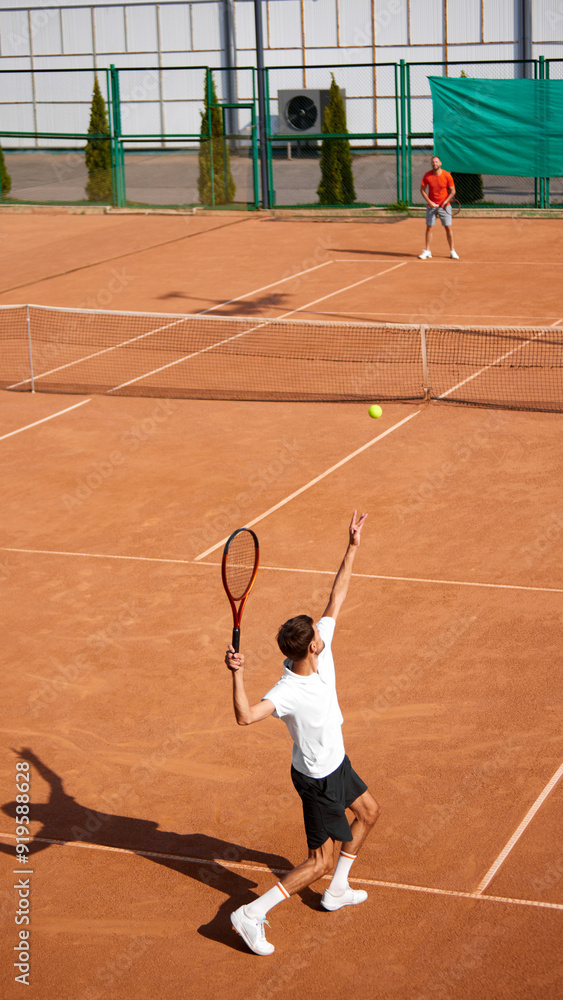 Vertical image of two men, tennis players on clay court playing, training, getting ready to serve and receive ball. Intense summer day game. Concept of sport, competition, active and healthy lifestyle Vertical image of two men, tennis players on clay court playing, training, getting ready to serve and receive ball. Intense summer day game. Concept of sport, competition, active and healthy lifestyle