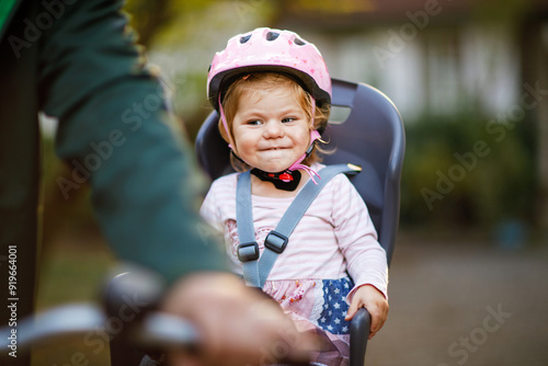 A smiling toddler girl wearing a pink helmet sits securely in a bicycle child seat, ready for a ride. The background is blurred, focusing on the child joyful expression