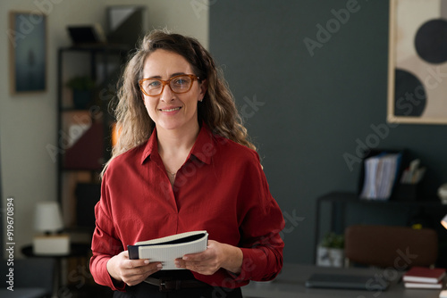 Slika na platnu Portrait of woman wearing glasses and holding open book while smiling