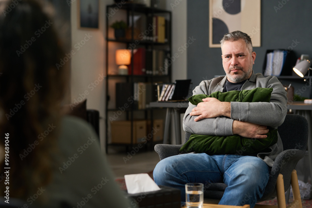 Man sitting in office holding green pillow with both hands, looking relaxed and thoughtful in well-decorated room Man sitting in office holding green pillow with both hands, looking relaxed and thoughtful in well-decorated room