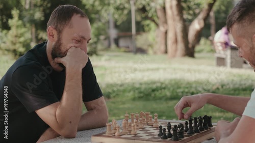 Two male friends are playing chess in the park on a sunny day. One is thinking hard about his next move, while the other makes his move. They are both smiling and having fun.