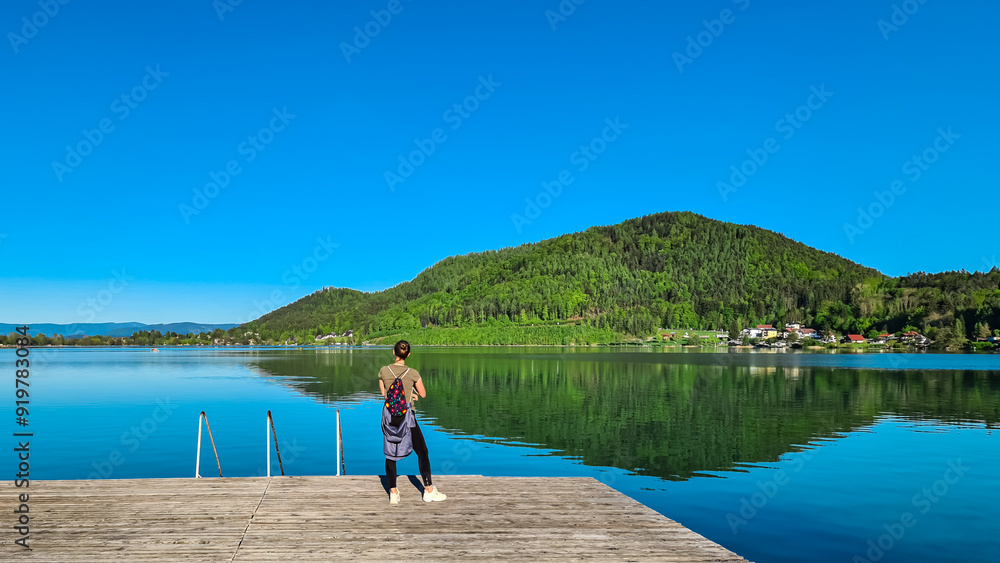 Woman at wooden pier looking at lake Klopein nestled amidst lush green forest in Carinthia, Austria. Crystal-clear water reflects vibrant blue sky. Rolling hills and mountains in Austrian Alps Woman at wooden pier looking at lake Klopein nestled amidst lush green forest in Carinthia, Austria. Crystal-clear water reflects vibrant blue sky. Rolling hills and mountains in Austrian Alps