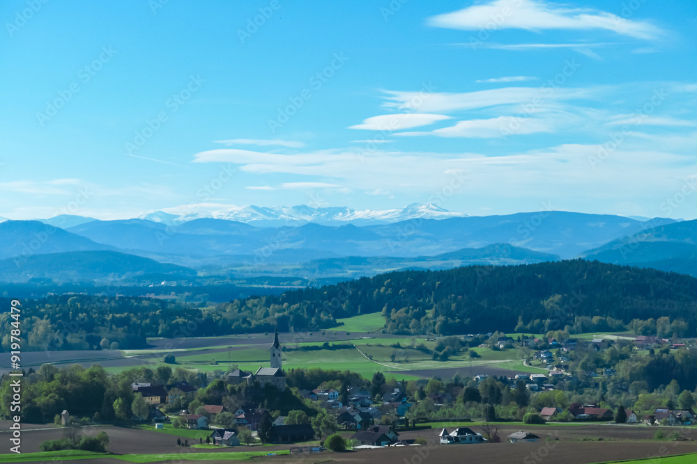 Fototapeta premium Alpine landscape with church in small village Stein im Jauntal nestled amidst rolling hills and lush greenery. Majestic mountain range with snow-capped peaks in Carinthia. Breathtaking backdrop