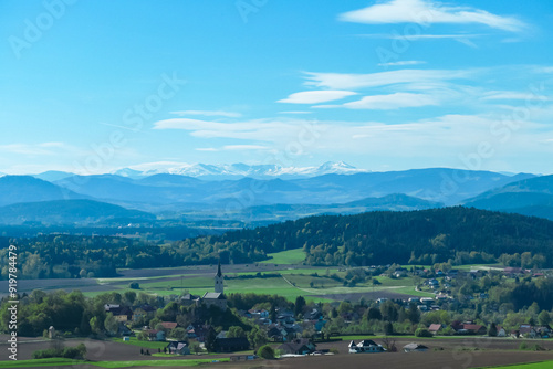 Alpine landscape with church in small village Stein im Jauntal nestled amidst rolling hills and lush greenery. Majestic mountain range with snow-capped peaks in Carinthia. Breathtaking backdrop photo