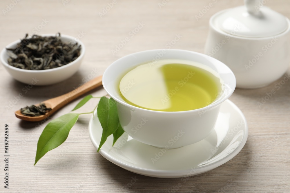 Refreshing green tea in cup, sugar bowl, spoon and leaves on wooden table, closeup