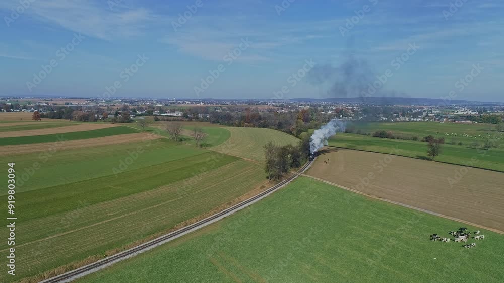 An Aerial of a Steam Train With Red Freight Cars Moves Through Vast, Patchwork Green And Brown Fields, Emitting White Smoke As It Winds Along A Rural Landscape, With A Town Visible In The Distant An Aerial of a Steam Train With Red Freight Cars Moves Through Vast, Patchwork Green And Brown Fields, Emitting White Smoke As It Winds Along A Rural Landscape, With A Town Visible In The Distant