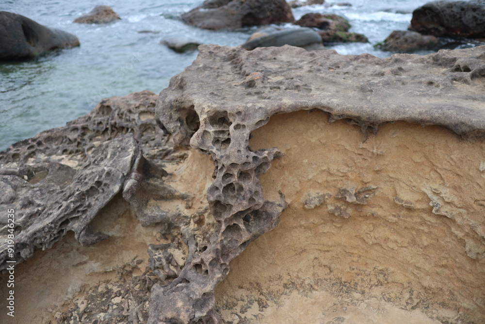 Fototapeta premium Close-up of a weathered and eroded rock formation on a coastline, showing intricate patterns and textures created by natural forces. The rock is a light brown color, with dark grey and black streaks.