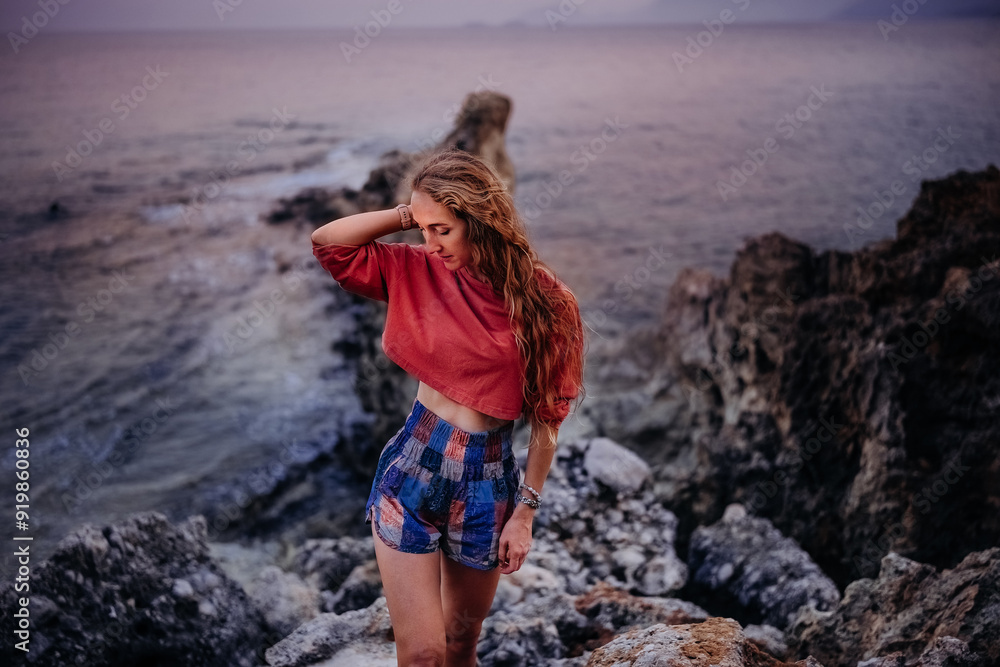Young woman in casual clothing standing on rocky beach at sunset near the ocean Young woman in casual clothing standing on rocky beach at sunset near the ocean