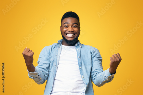 Portrait of overjoyed black man celebrating success with clenched fists and euphoric face expression on yellow studio background with copy space photo