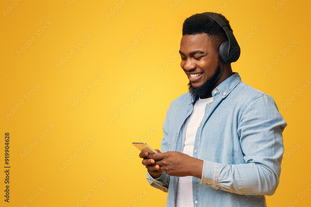 App for music. Cheerful african american guy in wireless headphones listening his favorite songs on smartphone, yellow background with free space App for music. Cheerful african american guy in wireless headphones listening his favorite songs on smartphone, yellow background with free space