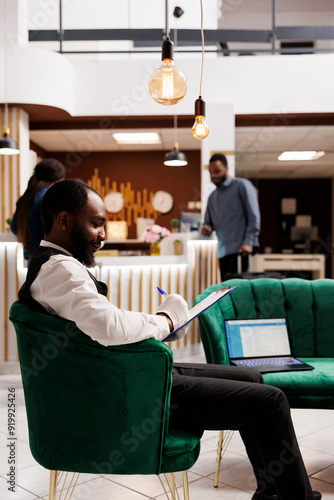 Фотография Young smiling African American hotel bellman filling errand card while sitting at lounge area