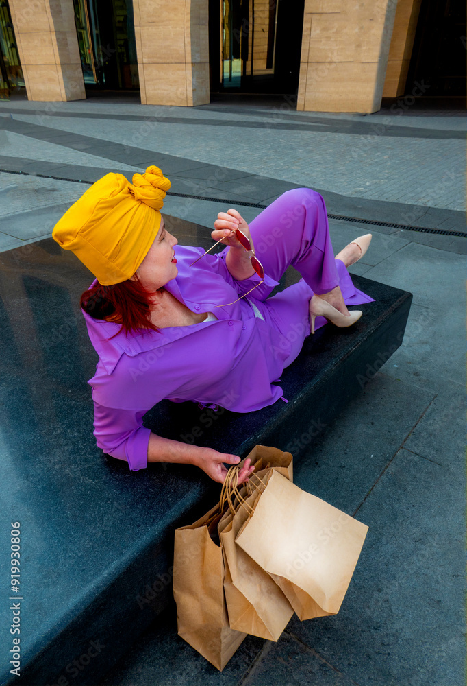 woman with craft shopping bags in the city. The concept of fashionable Consumerism and ecology. Adult Asian woman in a purple suit with red and yellow accents enjoys shopping. rest between purchases