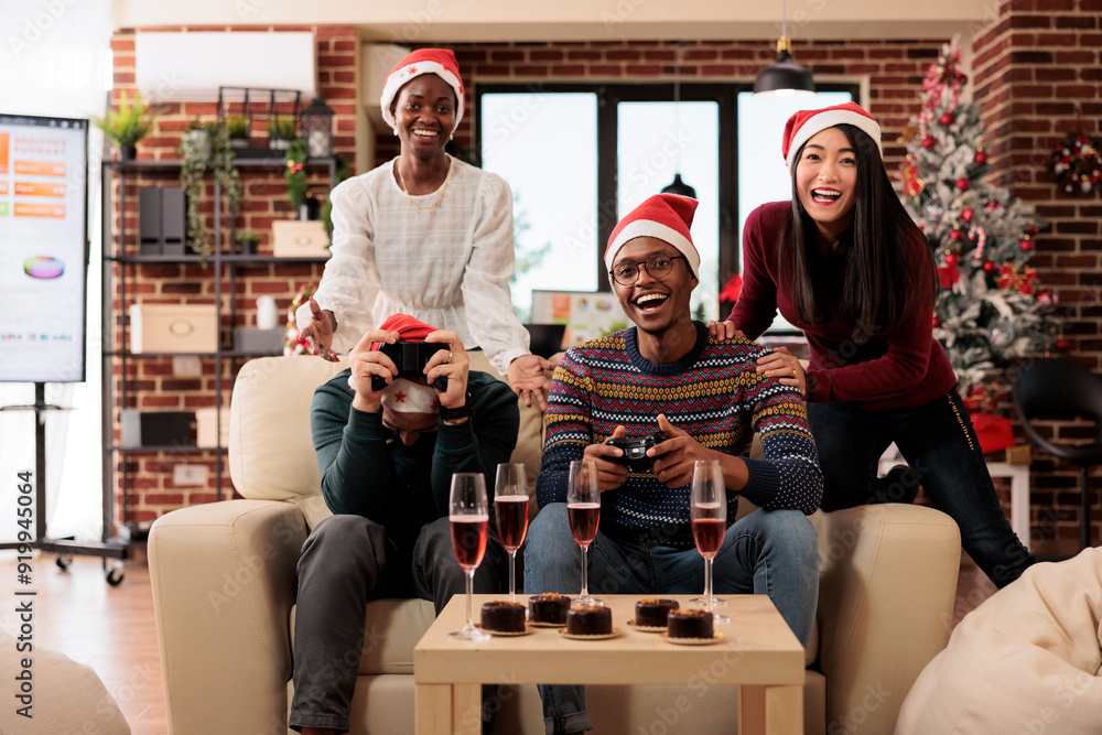 Naklejka premium Cheerful colleagues celebrating new year together, holding gamepads and enjoying holiday party in decorated office. Happy coworkers sitting on couch and playing videogame on christmas eve