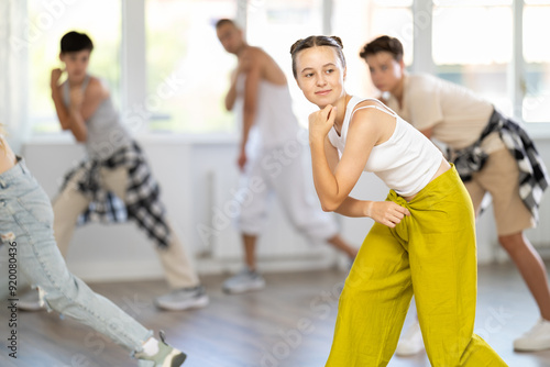 Young girl in bright clothes, together with group of teenage friends, learn to dance hip hop in choreographic hall. Students move to modern music, do moonwalking, squats, bends, work with body photo