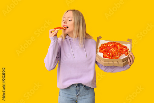 Young woman eating piece of tasty pizza and holding cardboard box on yellow background