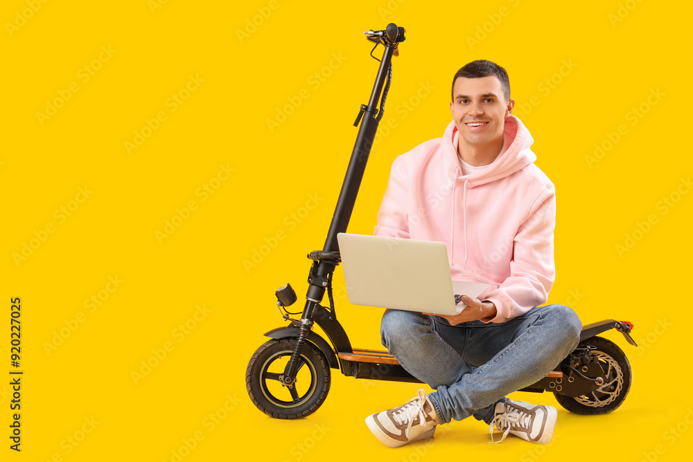Young man with electric scooter using laptop on yellow background Young man with electric scooter using laptop on yellow background