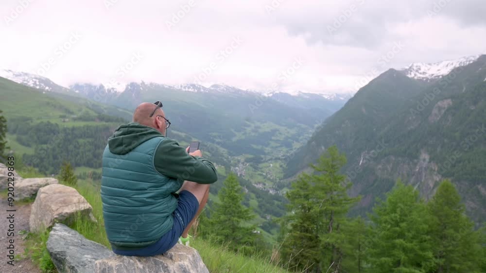 A man sitting on a rock in a mountainous landscape, enjoying the scenic view while using his smartphone. The serene setting is surrounded by lush greenery and distant snow-capped peaks. A man sitting on a rock in a mountainous landscape, enjoying the scenic view while using his smartphone. The serene setting is surrounded by lush greenery and distant snow-capped peaks.