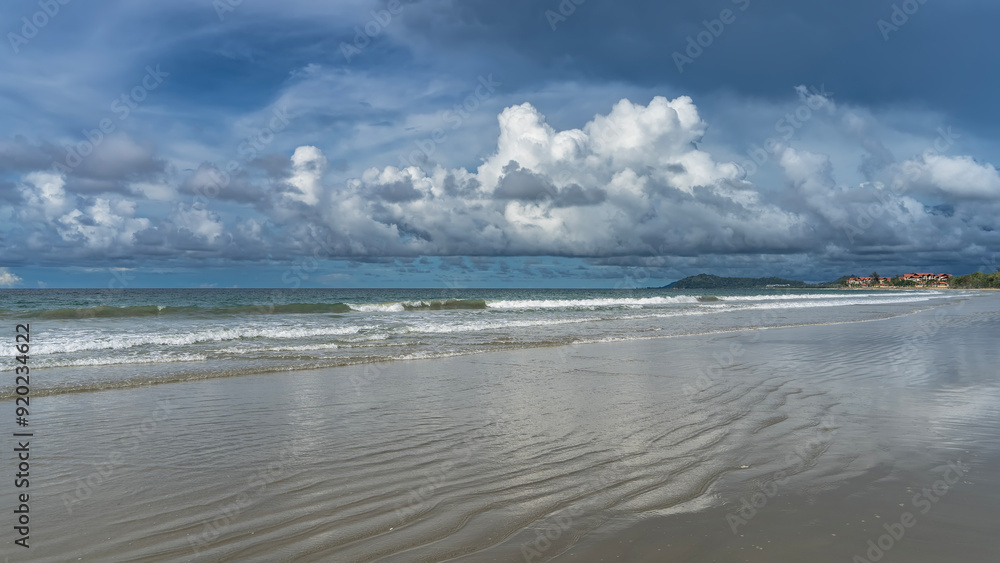 Beautiful seascape. The waves of the turquoise ocean roll, foaming and spreading over the deserted beach.  The hotel is far away. Clouds in the blue sky. Reflection on wet sand. Malaysia. Borneo.