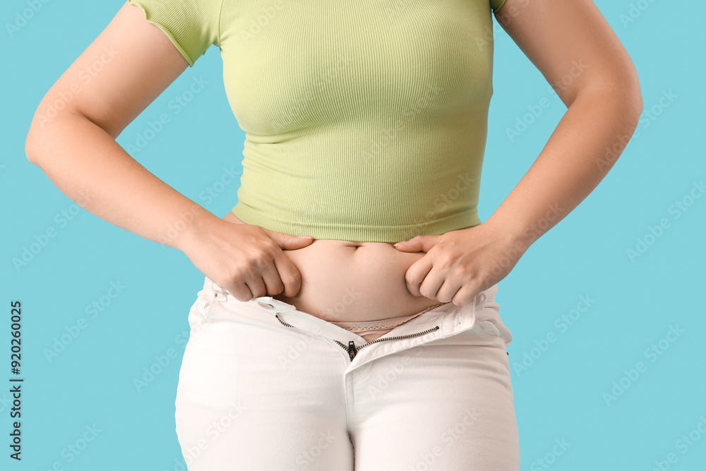 Young woman in tight jeans on blue background, closeup. Weight gain concept Young woman in tight jeans on blue background, closeup. Weight gain concept