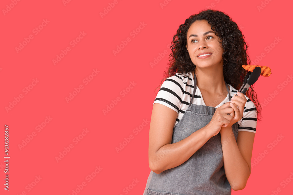 Beautiful young happy African-American woman with tasty grilled slice of carrot and tongs on red background Beautiful young happy African-American woman with tasty grilled slice of carrot and tongs on red background