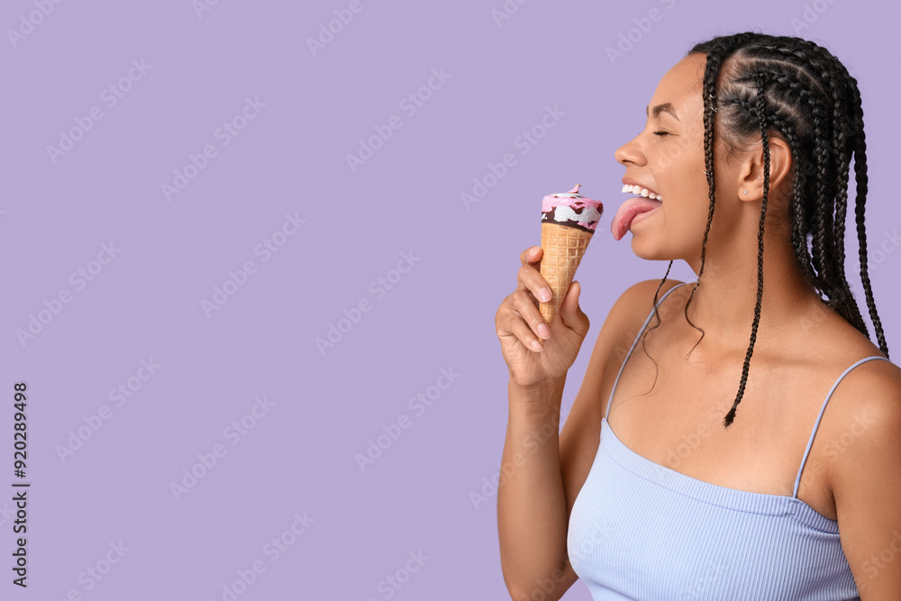 Beautiful young African-American woman eating sweet ice-cream in waffle cone on lilac background Beautiful young African-American woman eating sweet ice-cream in waffle cone on lilac background