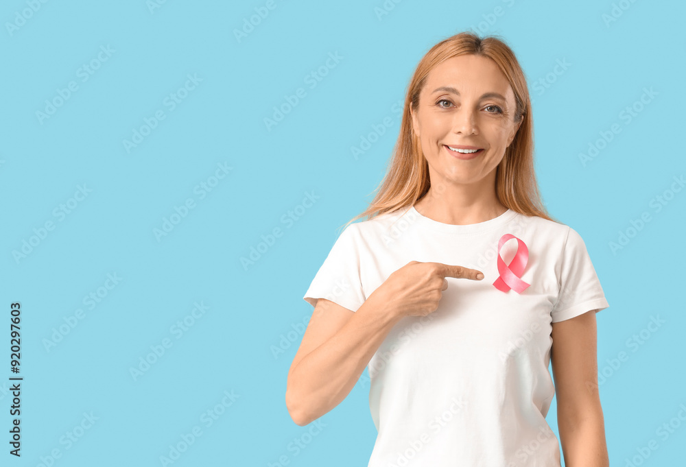 Mature woman pointing at pink ribbon on blue background. Breast cancer awareness concept Mature woman pointing at pink ribbon on blue background. Breast cancer awareness concept