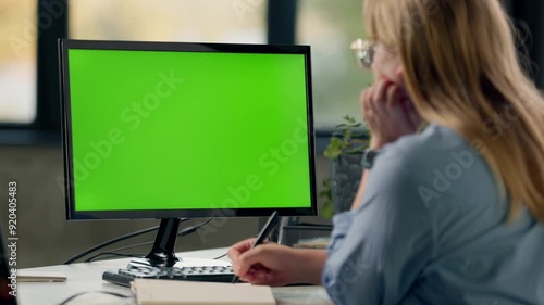 Young Woman Looks at Computer with Green Screen Mock Up Display and Writes Something in Notebook. Girl Working In Office On Computer With Green Screen