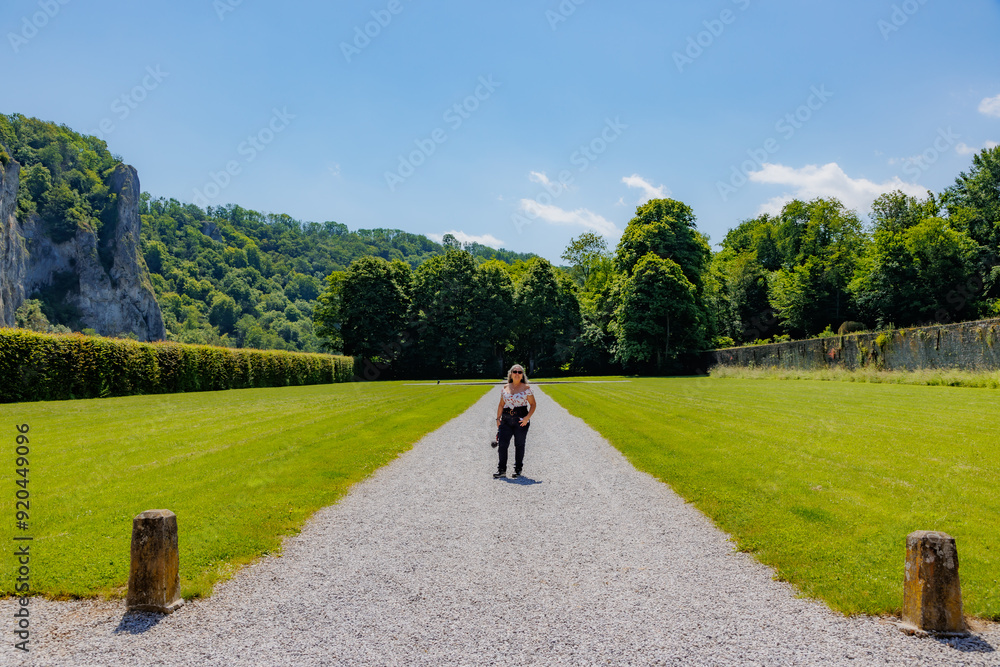 Gravel path with female tourist standing in garden outside Freyr Castle, lush trees and rocky mountain against blue sky in blurred background, sunny summer day in Namur, Wallonia, Belgium Gravel path with female tourist standing in garden outside Freyr Castle, lush trees and rocky mountain against blue sky in blurred background, sunny summer day in Namur, Wallonia, Belgium