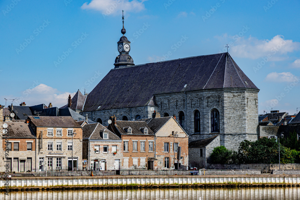 Naklejka premium Old buildings and back view of St. Hilarius church against blue sky in background, cityscape of Givet town, Meuse river in foreground, sunny summer day in Ardennes department, France