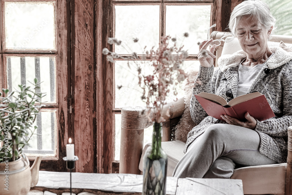 Old retired 70s lady reading book at home, resting on armchair close the window in cozy living room. Mature reader relaxing enjoying reading pleasure holding glasses in hand Old retired 70s lady reading book at home, resting on armchair close the window in cozy living room. Mature reader relaxing enjoying reading pleasure holding glasses in hand