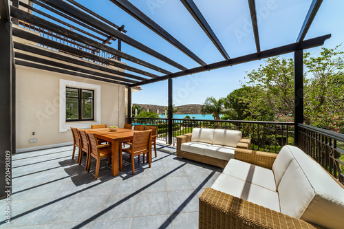 Balcony or terrace with outdoor furniture under wooden beams meant for roof, overlooking garden and sea view. Space combines comfort and natural beauty. Perfect for relaxation and enjoying.