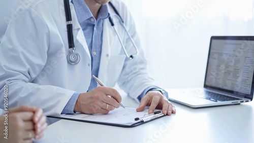 A doctor and a patient. The physician, wearing a white medical coat over a blue shirt, is filling out a medical record form during a consultation in the clinic. Medical service