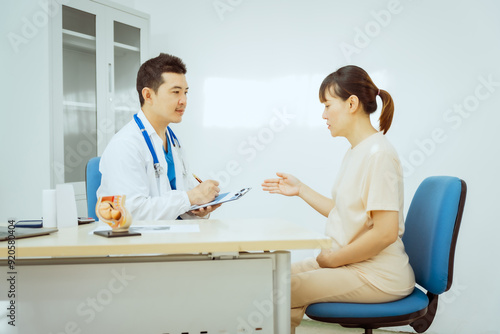 A male Asian pediatrician in a white lab coat sits at his desk, warmly greeting a middle-aged pregnant woman for a prenatal consultation, offering care and support in a clinical setting.