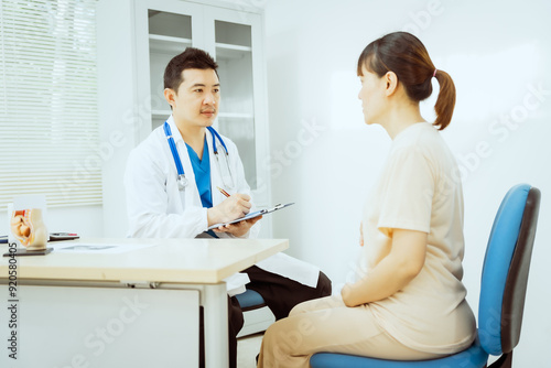 A male Asian pediatrician in a white lab coat sits at his desk, warmly greeting a middle-aged pregnant woman for a prenatal consultation, offering care and support in a clinical setting.