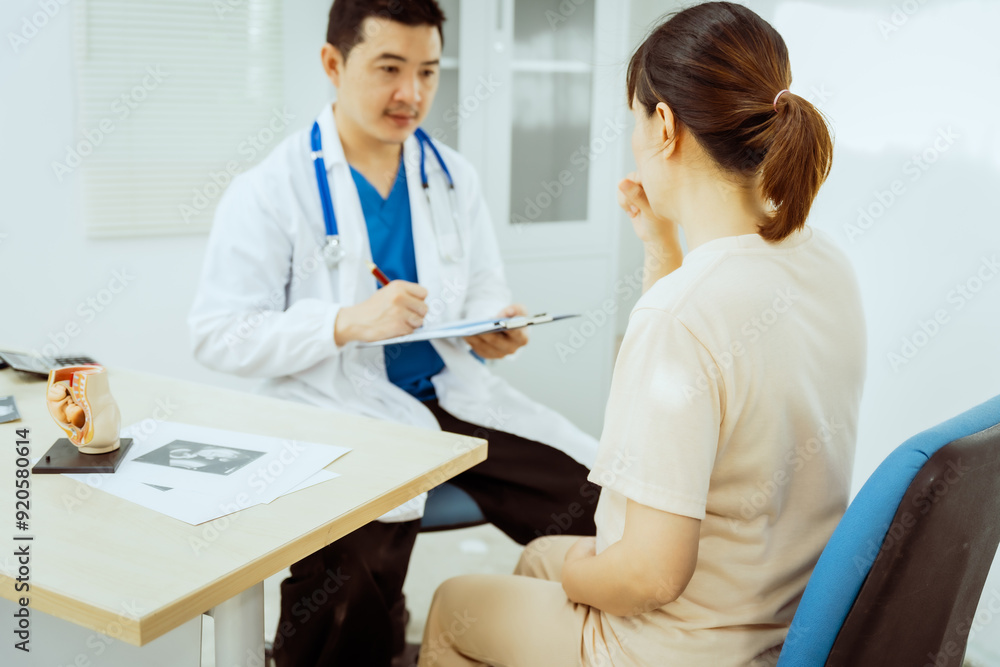 A male Asian pediatrician in a white lab coat sits at his desk, warmly greeting a middle-aged pregnant woman for a prenatal consultation, offering care and support in a clinical setting. A male Asian pediatrician in a white lab coat sits at his desk, warmly greeting a middle-aged pregnant woman for a prenatal consultation, offering care and support in a clinical setting.