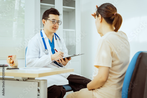 A male Asian pediatrician in a white lab coat sits at his desk, warmly greeting a middle-aged pregnant woman for a prenatal consultation, offering care and support in a clinical setting.