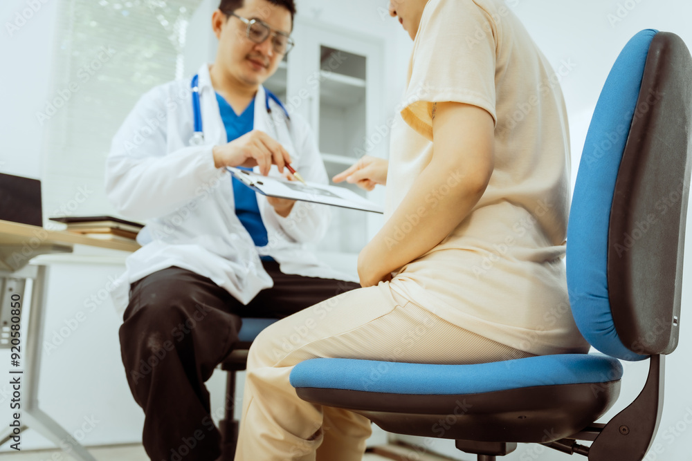 Obraz premium A male Asian pediatrician in a white lab coat sits at his desk, warmly greeting a middle-aged pregnant woman for a prenatal consultation, offering care and support in a clinical setting.
