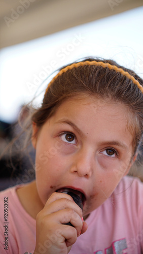 Close-up of a young girl eating an ice cream, her eyes looking upward, with a blurred background