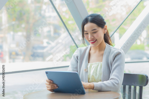Young asian professional business female in light gray long sleeve shirt is looking at tablet confidently in her hands while she is sitting working at coffee shop among business background in city