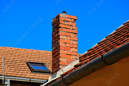 damaged clay brick chimney with weathered and spalling surface. clay tile roofing. gray metal flashing. strong shadow. glass skylight. blue sky background. building, home maintenance, upkeep concept