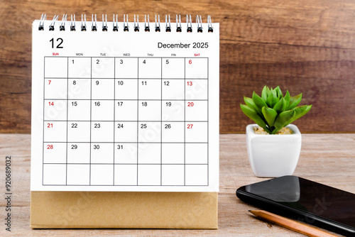December 2025 desk calendar on wooden table with black smartphone.
