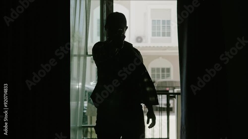 Silhouette of a man in a long-sleeved striped shirt and a cap, standing with a gun raised in front of him, looking menacing, at the entrance of the victim's house.
