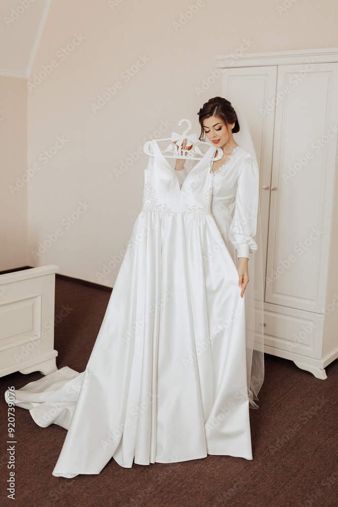 Fototapeta premium A bride is standing in front of a white dress hanging on a hanger. She is wearing a white veil and a necklace. The dress is long and elegant, and the bride is looking at it with a smile on her face